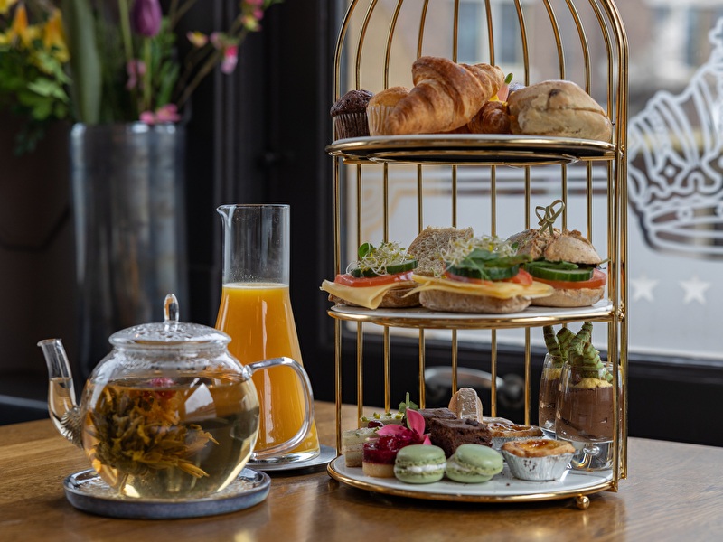 Een 3 laags etagere met daarop crossaints, belegde gezonde broodjes, macarons en cakjes en een glas jus de orange en een pot thee op tafel.