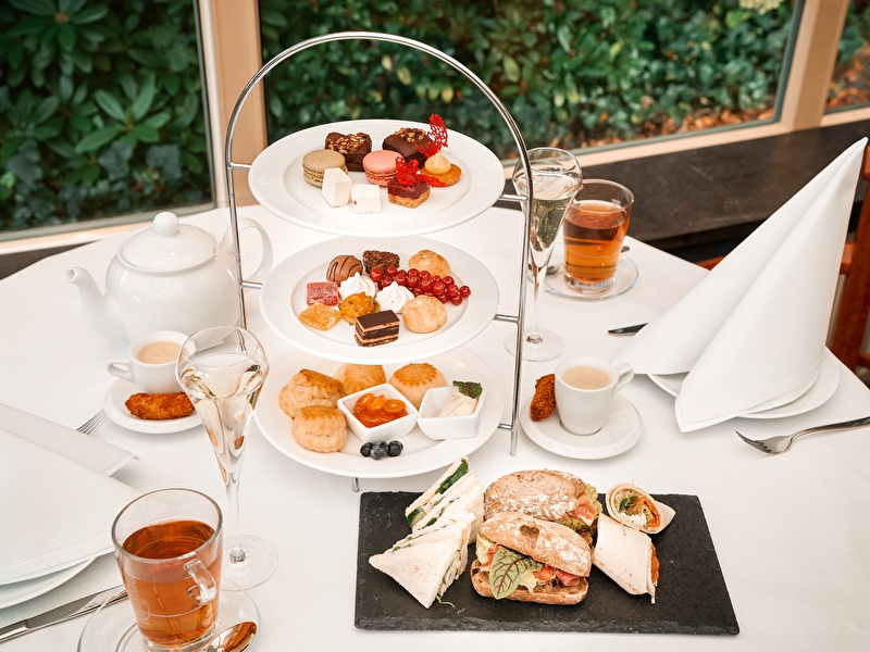 Een 3 laags etagere met daarop crossaints, belegde gezonde broodjes, macarons en cakjes en een glas jus de orange en een pot thee op tafel.