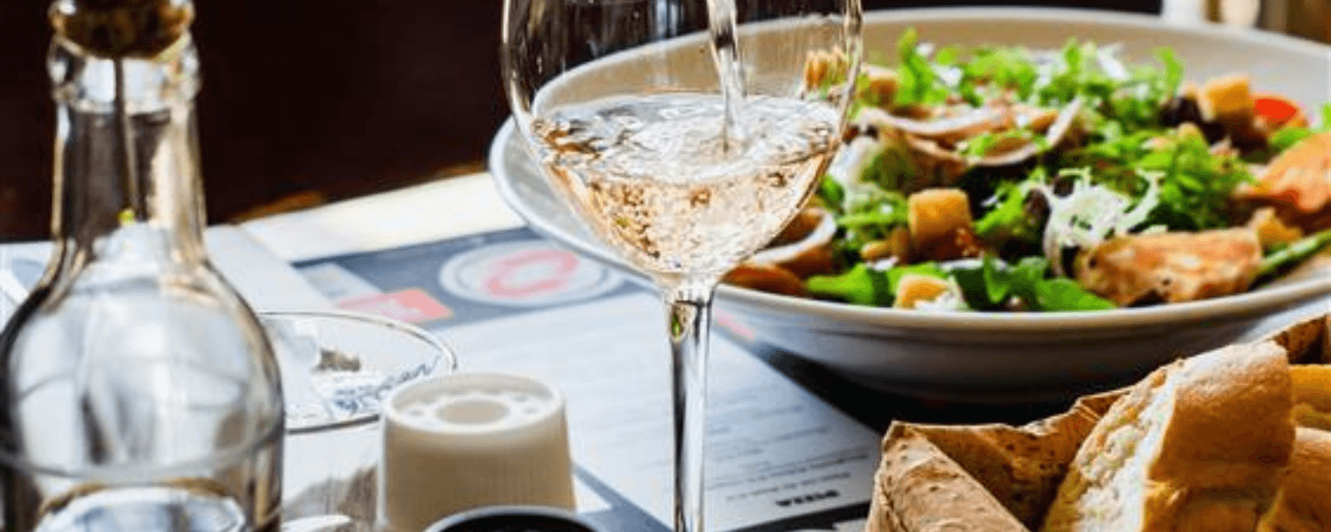 Een glas witte wijn wordt ingeschonken op een tafel in een restaurant, met een frisse salade en een mandje brood op de achtergrond. De tafel is sfeervol gedekt met olijfolie, peper en zout.