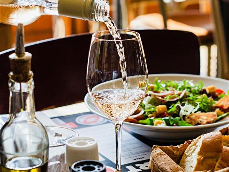 Een glas witte wijn wordt ingeschonken op een tafel in een restaurant, met een frisse salade en een mandje brood op de achtergrond. De tafel is sfeervol gedekt met olijfolie, peper en zout.