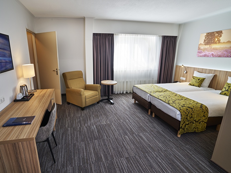 Superior kamer in Amrâth Hotel Lapershoek Arenapark Hilversum met een tweepersoons bed op het bed ligt een geel met zwart geprint sprei met twee bijpassende kussentjes en in de hoek van de kamer staat een bureau en een stoel