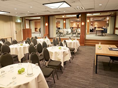 Amrâth Hotel Lapershoek Arenapark, vergaderzaal Lapers, Bach en Beethoven gecombineerd met ronde tafels met een wit kleed en zwarte stoelen ernaast. Vooraan in de ruimte staat een centrale tafel met een stoel.