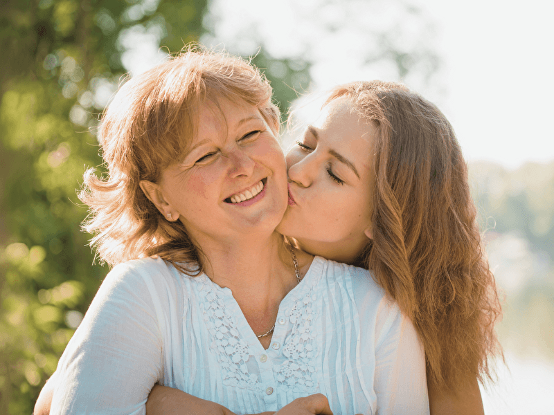 Laughing mother is lovingly embraced and kissed on the cheek by her daughter during a sunny day in a green, natural setting.